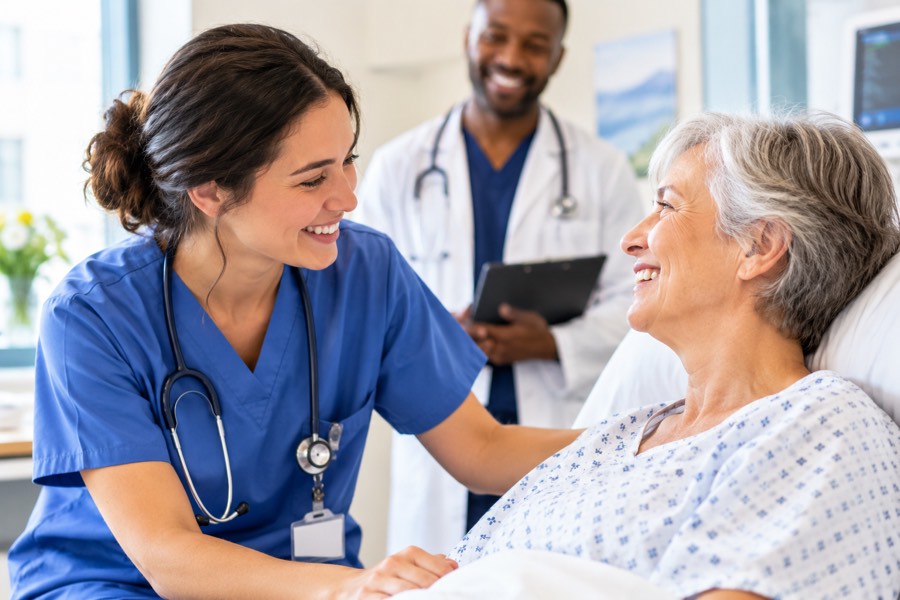 A nurse sitting with an older patient on a ward, a doctor with a clipboard behind them